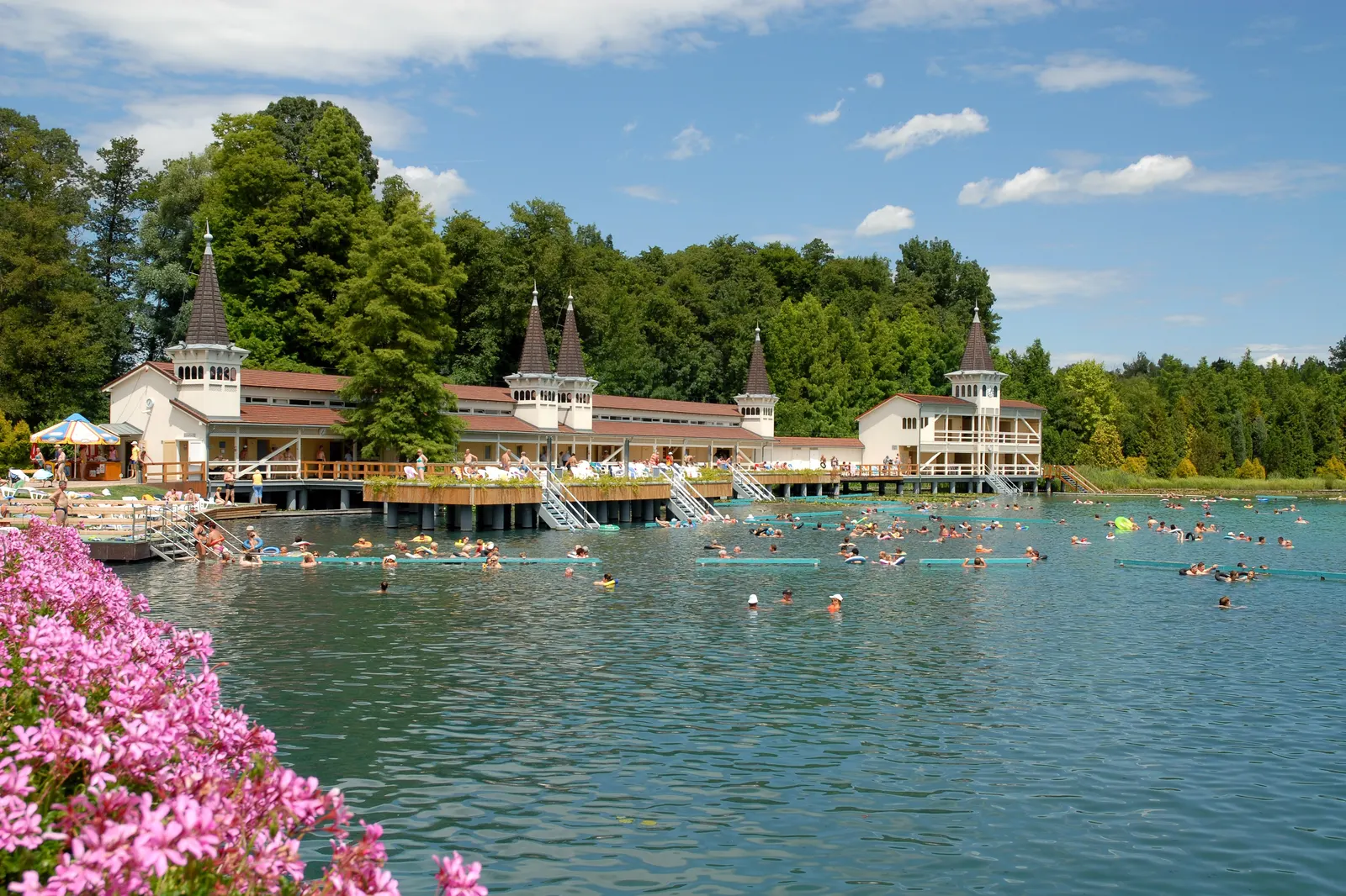 Lac thermal de Hévíz vu depuis la rive, pavillon en bois aux tourelles caractéristiques, baigneurs dans l'eau turquoise entourée de verdure